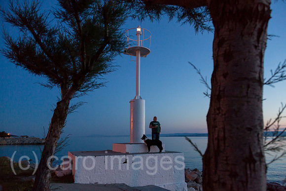 Kroatien, Krk (Hauptstadt der Insel Krk) - Mann mit Hund am Leuchtturm des Yachthafens in der Abenddaemmerung
